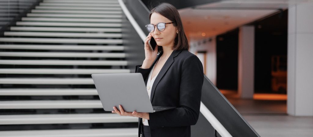 business woman in a formal suit with a laptop in the office of the business center stands on the steps in the lobby and works online. Work in a corporation as a marketing financier or lawyer. a successful woman goes to negotiations